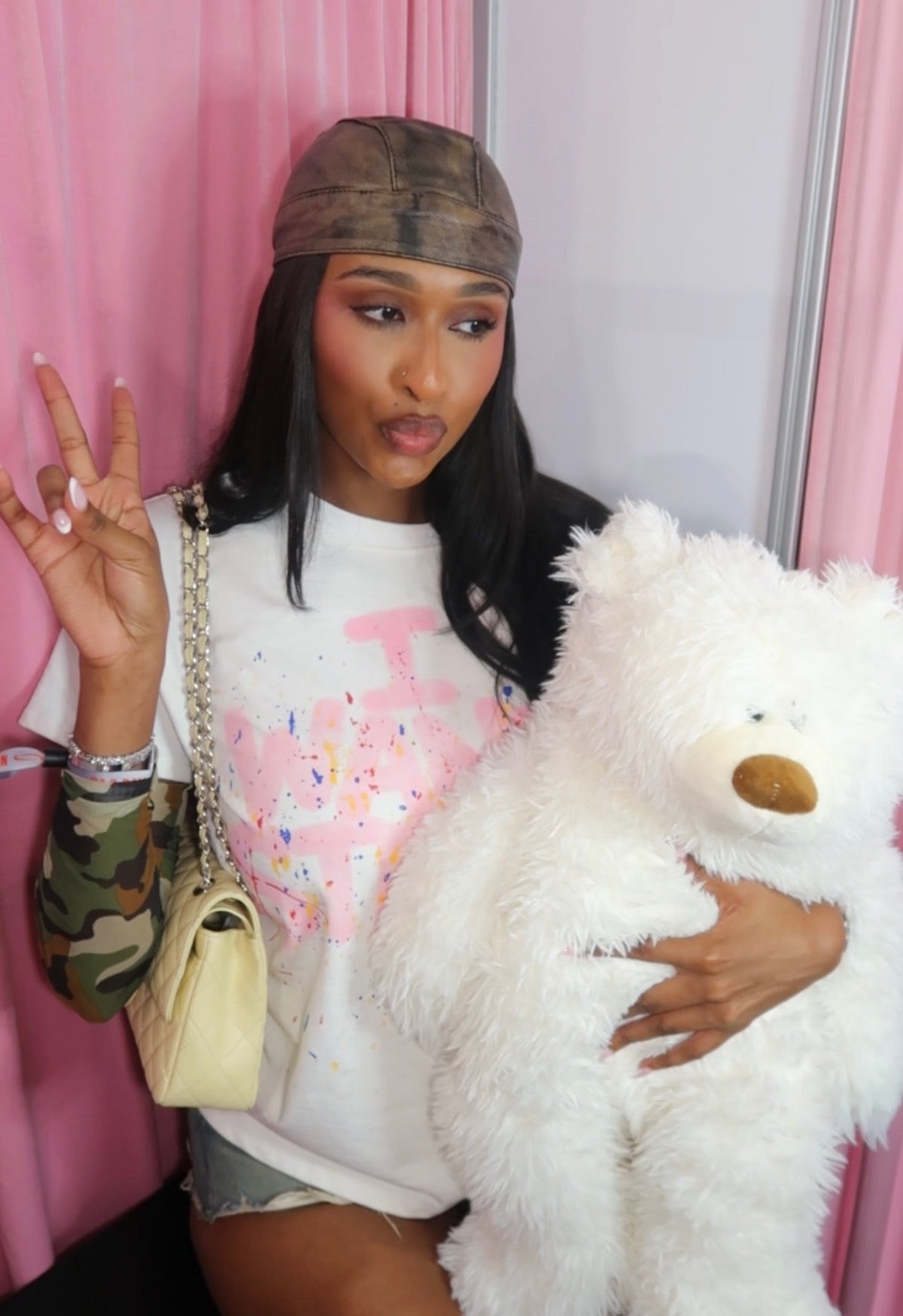 A woman posing in a pink room holding a large white teddy bear, wearing a camouflage cap, a white t-shirt with a pink paint splatter design, and carrying a small beige shoulder bag.